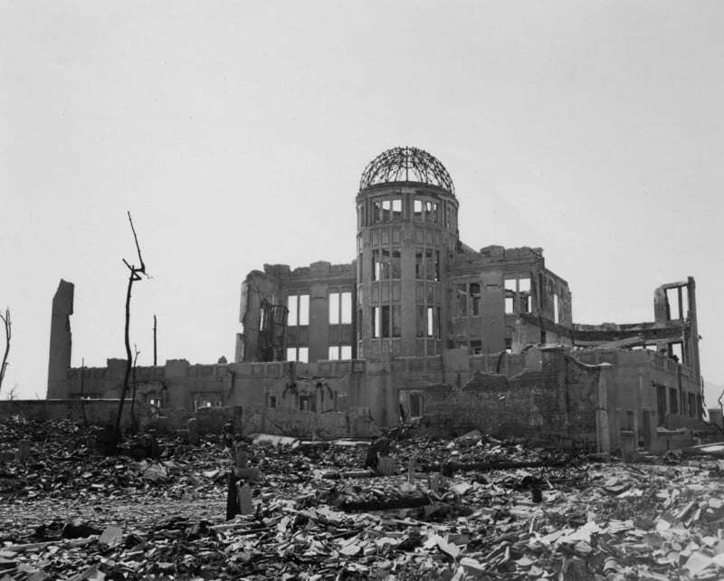 The ruins of the Hiroshima Prefectural Industrial Promotion Hall, now known as the Atomic Bomb Dome, stand amid debris and destruction after the atomic bombing in 1945. Two bare trees are visible in the foreground.