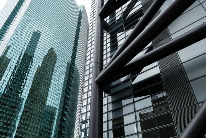 Glass skyscrapers with reflective windows, showing nearby buildings mirrored in the glass. Large black structural beams cross the foreground, adding contrast to the modern urban architecture.