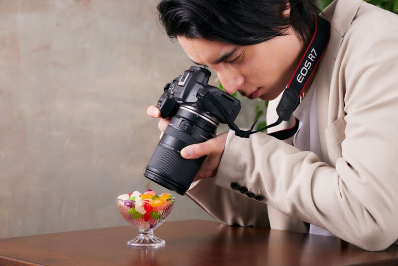 A person in a beige jacket is using a DSLR camera to photograph a glass bowl filled with colorful candy on a wooden table. The camera strap has "EOS R" written on it.