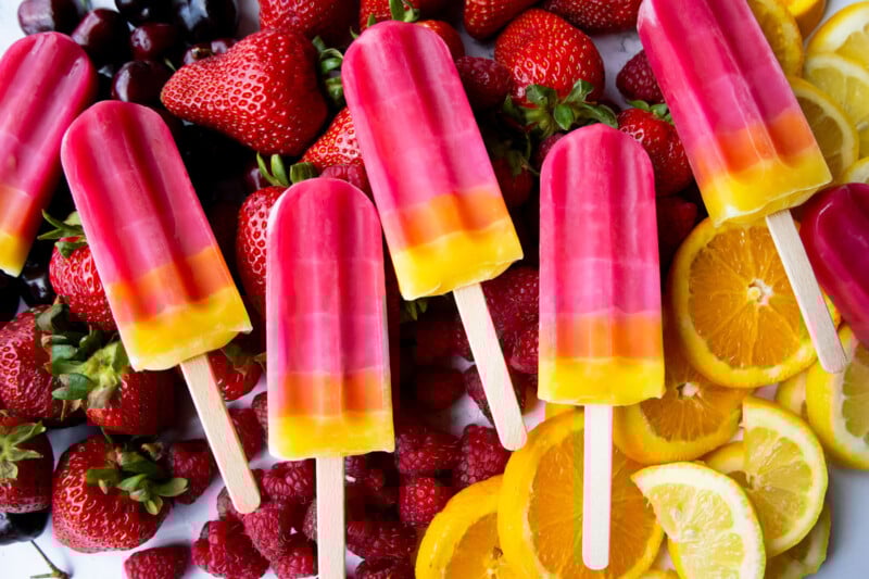 Colorful layered popsicles in pink, orange, and yellow rest on a bed of fresh strawberries, raspberries, cherries, orange slices, and lemon slices. The scene is vibrant and summery.