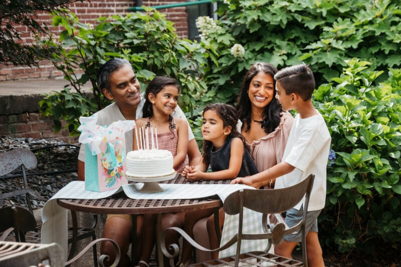 A family of five sits around a table outdoors, smiling at each other. A birthday cake with candles and a colorful gift bag are on the table. Greenery and flowers are in the background.