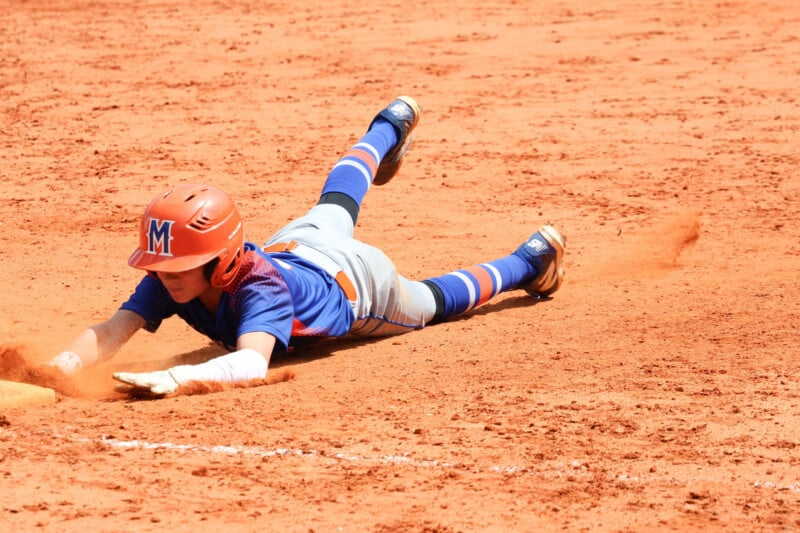 A young baseball player in a blue and orange uniform slides headfirst into a base on a dirt field, kicking up dust with an outstretched arm reaching for the base.