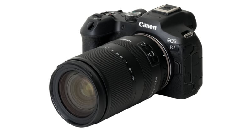 A black Canon EOS R7 digital camera with an attached zoom lens is angled to show the front and right side, displaying buttons, dials, and the Canon logo.