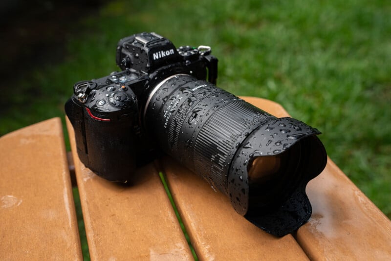 A Nikon camera with a large zoom lens, covered in raindrops, sits on a wooden surface outdoors with green grass in the background.