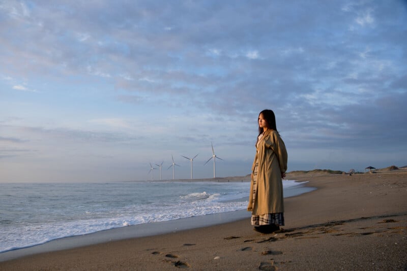 A woman in a long coat stands on a sandy beach near the shoreline, looking out at the sea. Wind turbines line the distant coast under a partly cloudy sky, and footsteps are visible in the sand.