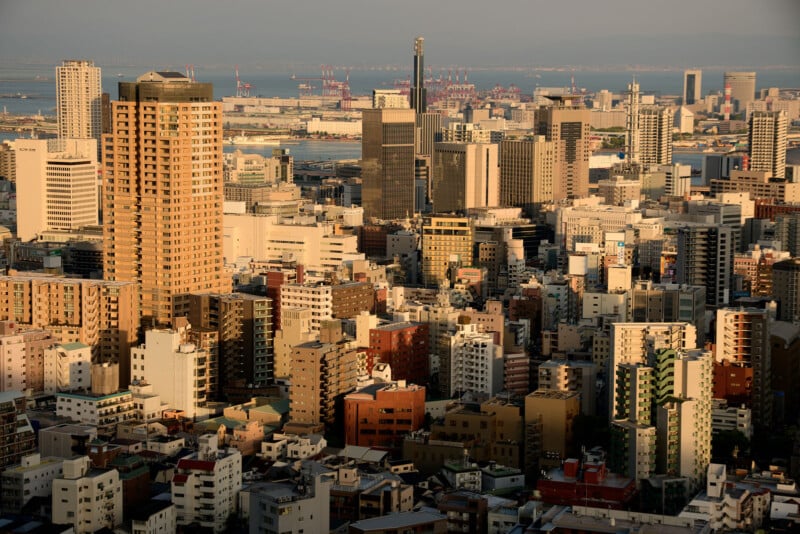 A dense urban skyline with many high-rise buildings, viewed in warm evening light, with an industrial port and water visible in the background.