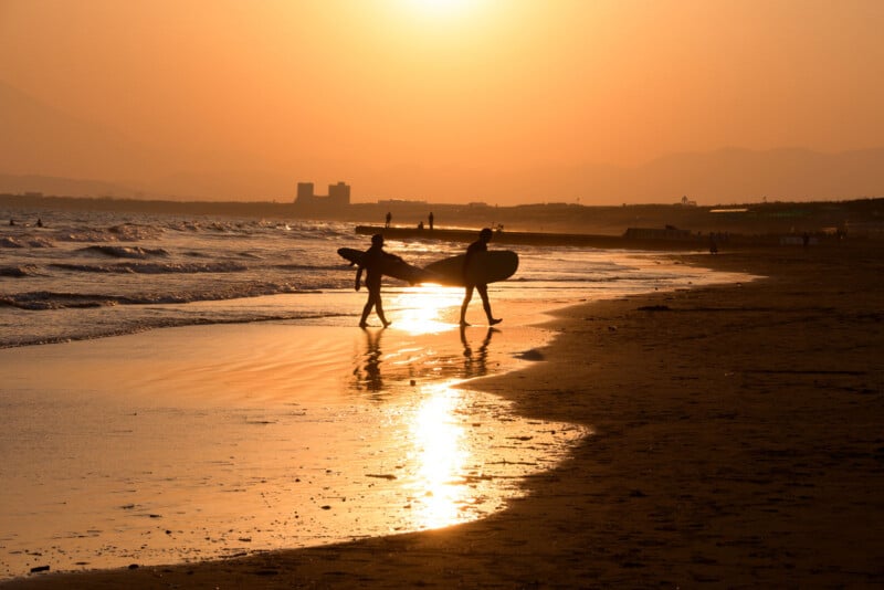 Two surfers carrying surfboards walk along a beach at sunset, with golden sunlight reflecting on the wet sand and gentle waves in the background.