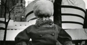 A person in a thick, high-collared coat and large, fluffy hat stands outside in winter, with the circular architecture of the Guggenheim Museum and bare trees in the background.