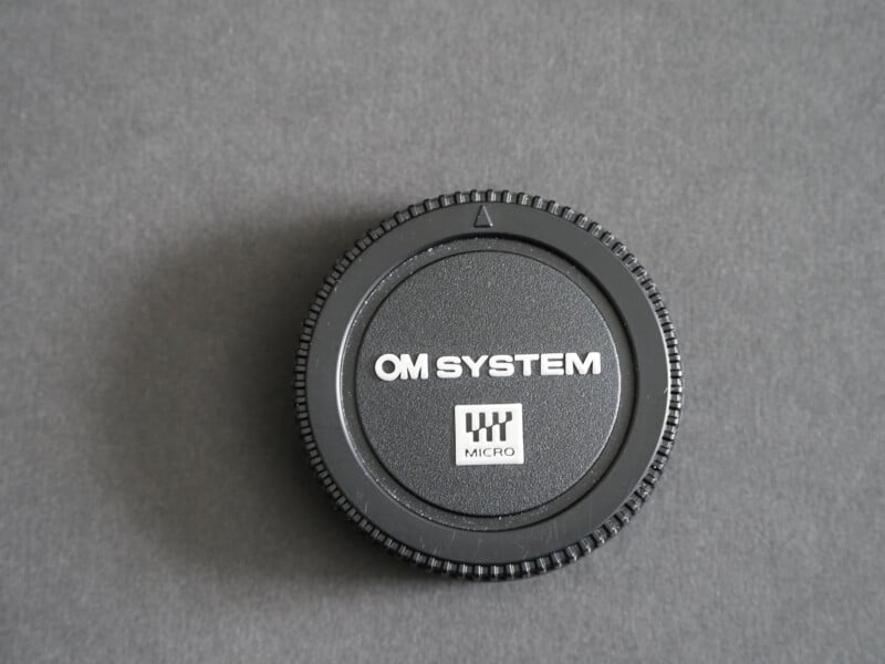 A black OM SYSTEM Micro camera body cap with white text is placed on a plain, dark gray surface.