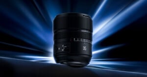 A black camera lens labeled “35” is displayed against a dramatic blue and black background with bright light streaks radiating outward. The lens sits on a dark, smooth surface.