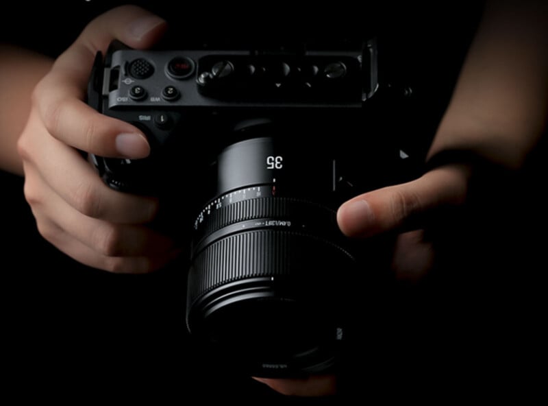 Close-up of two hands holding a black digital camera with a 35mm lens, against a dark background. The camera is centrally positioned and the hands are adjusting the lens or preparing to take a photo.