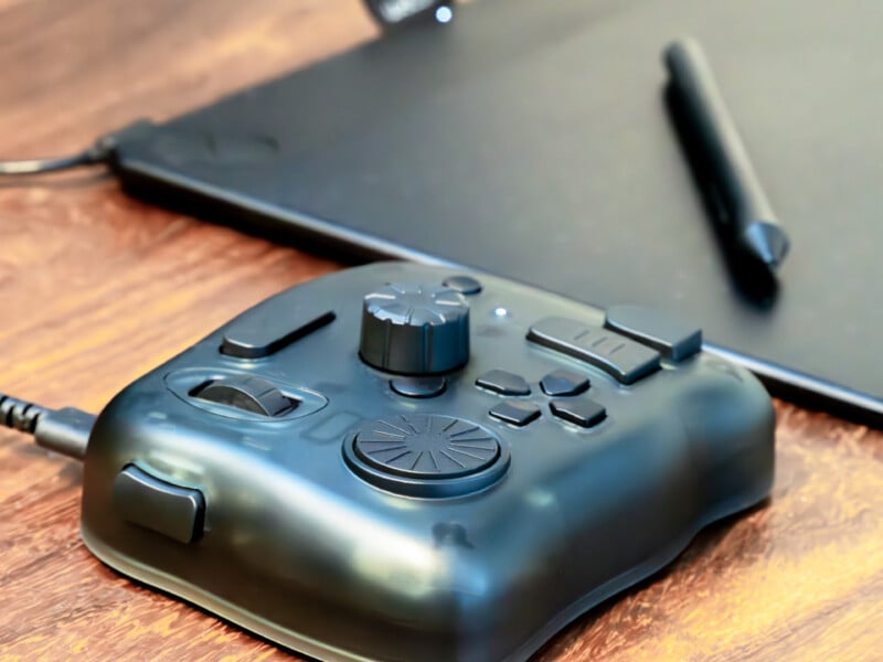 A close-up of a black digital editing controller with multiple buttons and dials, placed next to a stylus and a graphics tablet on a wooden surface.