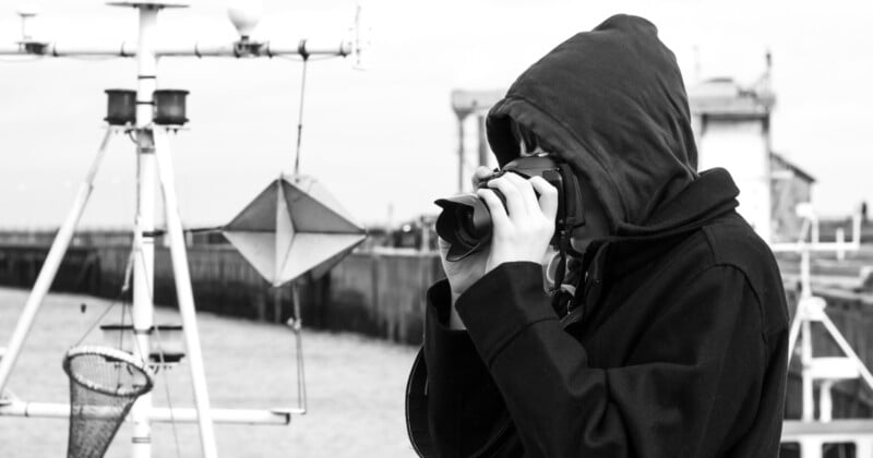 A person in a dark hooded coat takes a photo with a camera near a waterfront, surrounded by weather instruments and nautical equipment. The background shows a blurred building and railing. The image is in black and white.