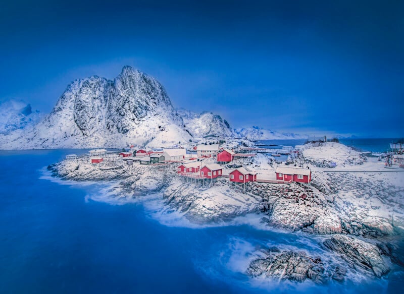 A snowy coastal village with red cabins sits on rocky shores beside calm blue water, surrounded by snow-covered mountains under a clear blue sky.