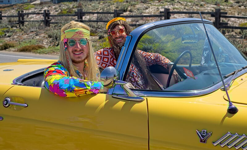 Two people dressed in colorful, retro hippie outfits sit in a yellow vintage convertible car with the top down, smiling and enjoying a sunny day. The background includes a wooden fence and greenery.
