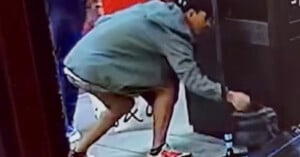 A person wearing a gray hoodie and red sneakers is bending down on a sidewalk, reaching toward a dark bag or object near a doorway.