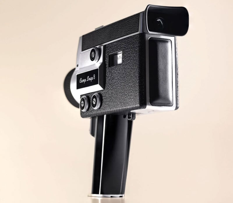 A vintage black and silver Super 8 camera with a textured grip and side markings is shown against a plain beige background. The camera is angled upward, highlighting its retro design and lens details.