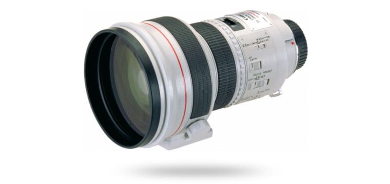 A large, white telephoto camera lens with multiple adjustment rings and switches, shown against a white background with a subtle shadow below it.