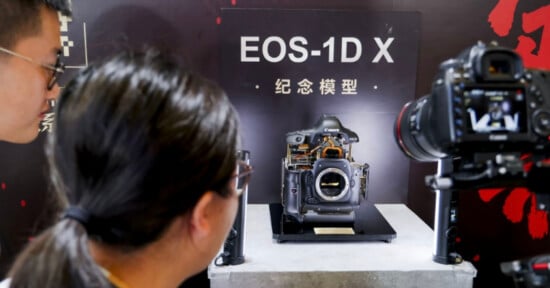 Two people look at a Canon EOS-1D X camera model displayed in a case, with a sign behind it and a camera on a tripod in the foreground recording or photographing the display.