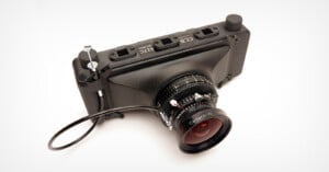A black, compact digital camera with several labeled ports on top and a wide-angle lens attached; a cable release is connected to the camera. The camera is photographed on a white background.