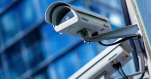 Two security cameras mounted on a wall, angled in different directions, with a modern glass building featuring blue windows blurred in the background.