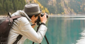 A person wearing a gray hat and jacket takes a photo with a camera on a tripod beside a calm lake, with trees and mountains in the background.