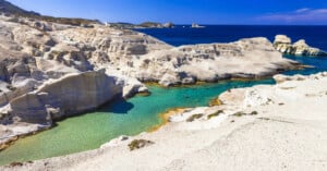 Rocky white cliffs surround a turquoise lagoon, with scattered people exploring the landscape. The sea and distant islands are visible under a clear blue sky in the background.