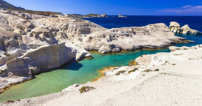 Rocky white cliffs surround a turquoise lagoon, with scattered people exploring the landscape. The sea and distant islands are visible under a clear blue sky in the background.