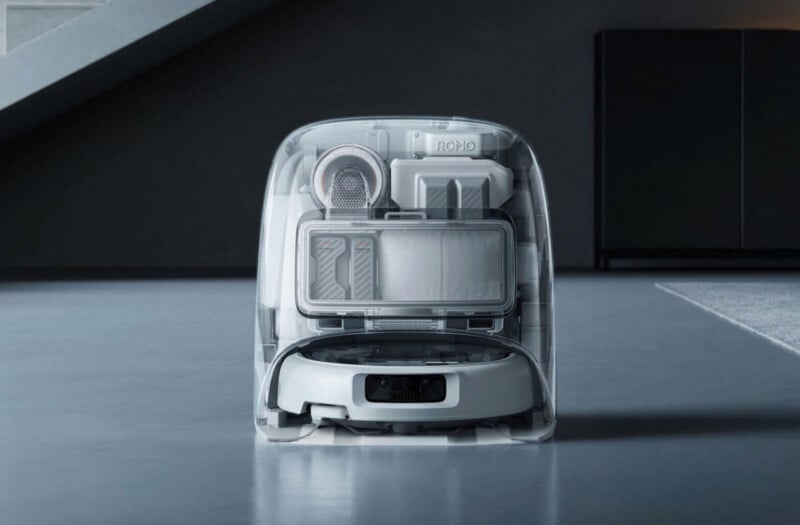 A robotic vacuum cleaner is docked inside a transparent charging station, revealing its internal components. The device is placed on a smooth, gray floor in a modern room with minimalistic decor.