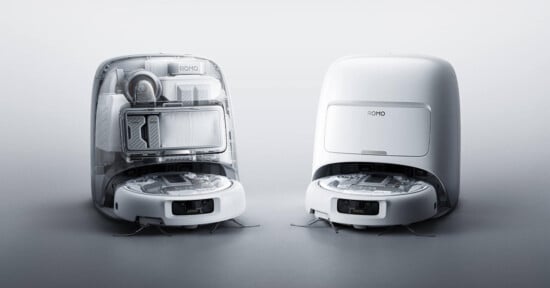 Two white robotic vacuum cleaners are shown side by side; one has its top open to reveal internal components and storage, while the other is closed, both set against a plain light background.