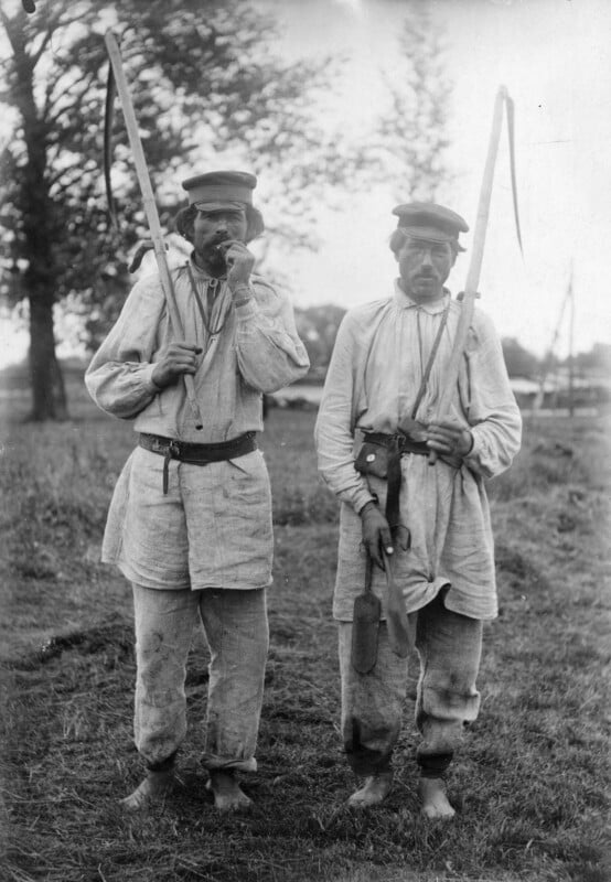 Two men in traditional rural clothing stand barefoot in a grassy field, each holding a scythe over their shoulder. They wear loose shirts, belts, and caps, with trees and blurry landscape in the background.