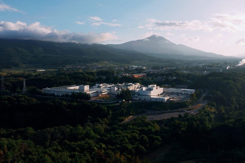 A large white industrial building surrounded by dense green trees, with a town and misty mountains in the background under a partly cloudy sky.