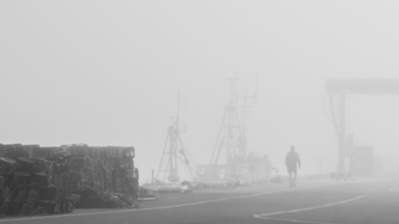 A person walks along a foggy dock, with fishing equipment and stacked crates barely visible in the thick mist.