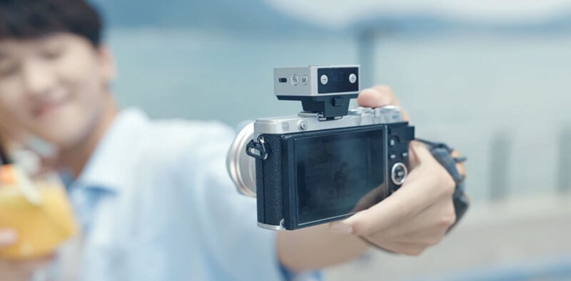 A person holds a digital camera at arm’s length, taking a selfie outdoors near water. The camera is in focus, showing its screen and controls, while the person in the background is blurred and smiling.