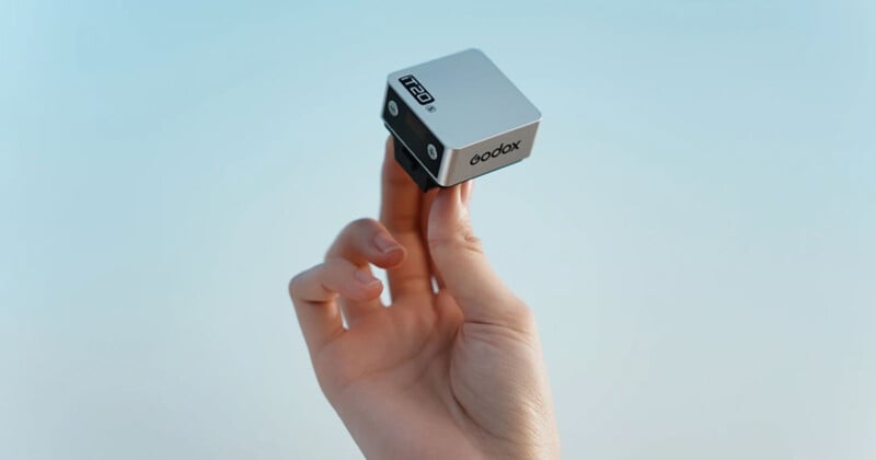 A hand holds a small, silver and black cubic device labeled "qodax" and "F20" between two fingers against a light blue background.