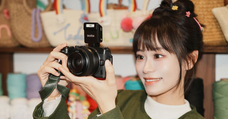 A young woman with a cheerful smile holds a camera with a flash attachment, aiming it forward. Colorful bags with rainbow handles are visible on shelves in the background.