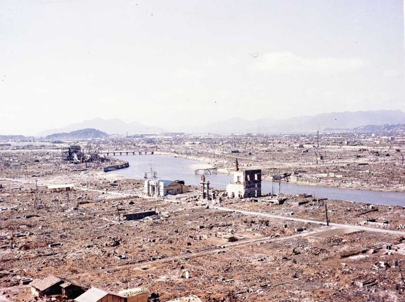 A wide view of a city devastated by destruction, with collapsed buildings and rubble along a river. A few structures remain partially standing, and a bridge crosses the river in the background under a hazy sky.