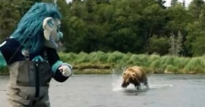 A person in a blue troll costume stands in shallow water, looking at a brown bear running through the water nearby. Green trees and grass line the background.