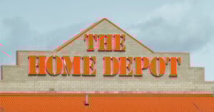 The Home Depot store exterior with large orange letters spelling "The Home Depot" on a beige brick wall and an orange roof below, set against a cloudy sky.
