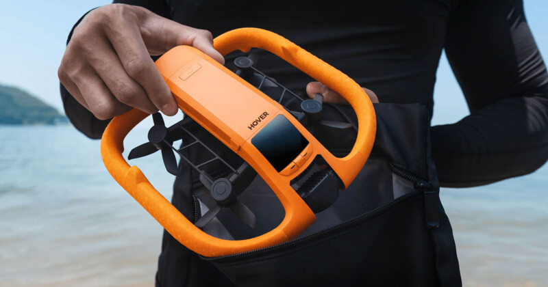 A person holding an orange Hover drone with protective guards, partially placing it into a black bag near a body of water with a blurred shoreline in the background.