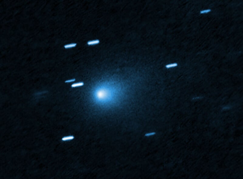 A bright, glowing blue comet with a faint tail appears in the center, surrounded by several streaks of light on a dark, star-filled background. The streaks suggest the motion of stars in the image.