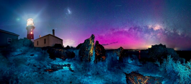 A lighthouse glows beside a small house on rocky terrain under a vibrant night sky, illuminated with stars, the Milky Way, and purple-blue auroras. The rocks and ground reflect blue and orange hues.