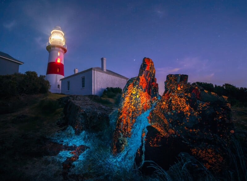 A brightly lit lighthouse stands behind a white building at dusk. In the foreground, rocky stones glow with blue and orange bioluminescence, creating a mystical effect under a starry sky.