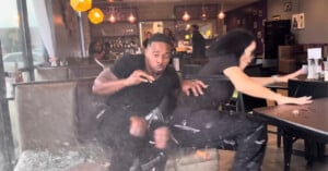 Two people in a restaurant booth react with shock as glass shatters and debris flies around them, indicating a sudden, dramatic event inside the busy dining area.