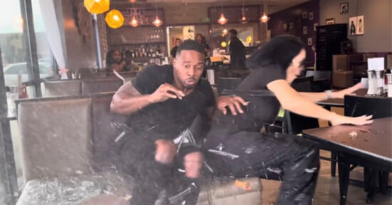 Two people in a restaurant booth react with shock as glass shatters and debris flies around them, indicating a sudden, dramatic event inside the busy dining area.