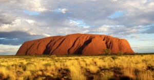 A large, reddish sandstone rock formation rises from a flat, grassy plain under a partly cloudy sky. This is Uluru in Australia, surrounded by dry grass and sparse vegetation.