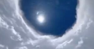 A circular opening in thick clouds reveals a bright sun shining in a deep blue sky, creating a dramatic contrast between the white clouds and the dark sky above.