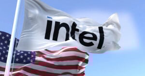 A white flag with the black Intel logo waves in the wind next to an American flag against a blue sky with clouds.