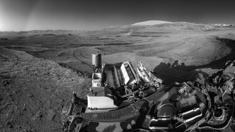 Black and white image showing the Mars Curiosity rover in the foreground with its equipment visible, overlooking a rocky, barren Martian landscape with hills and layered terrain in the distance under a clear sky.