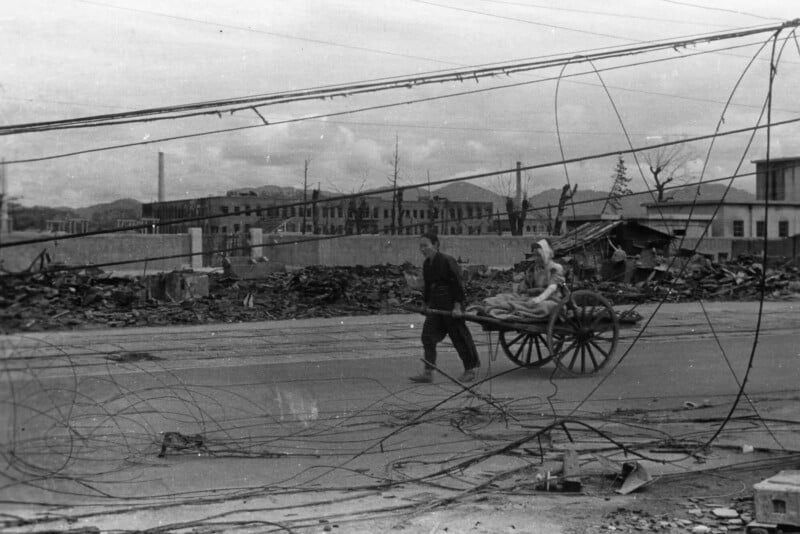 A person pulls a cart carrying another person through a street filled with debris and downed wires, with damaged buildings and rubble visible in the background. The scene looks desolate and war-torn.